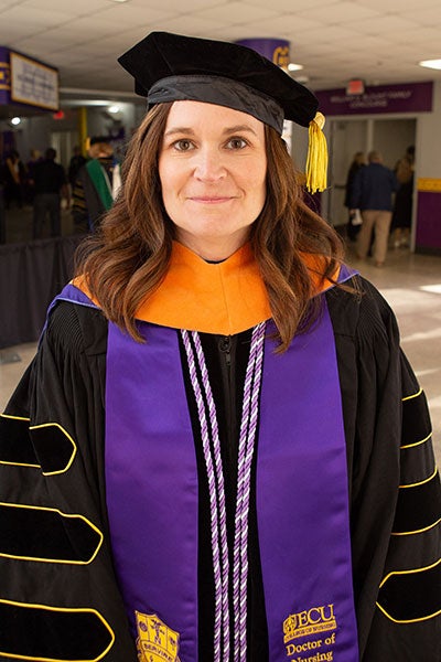 : A woman with long brown hair wearing academic regalia poses for a photo indoors.