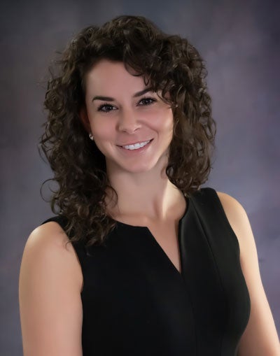 Professional headshot of a woman with curly dark hair, wearing a sleeveless black dress and smiling at the camera against a dark studio background.