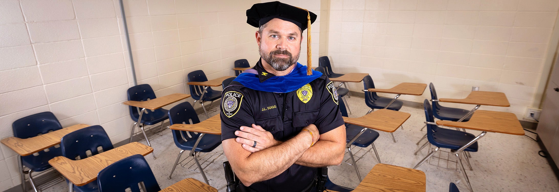 A man in a police uniform and a graduation cap with gold tassel stands with arms crossed in a classroom with desks.