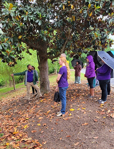 A bearded man in khaki pants and a rain jacket points toward a culvert while speaking with several faculty members wearing jackets and carrying umbrellas.