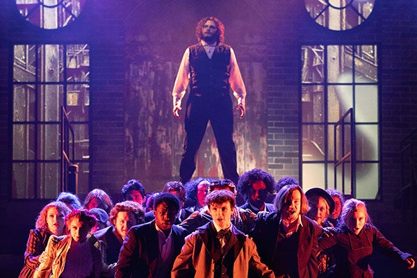 A man wearing a black vest with a white long sleeve shirt and black pants seemingly stands atop a group of people wearing nineteenth century clothing on a stage with two large windows in the background. 