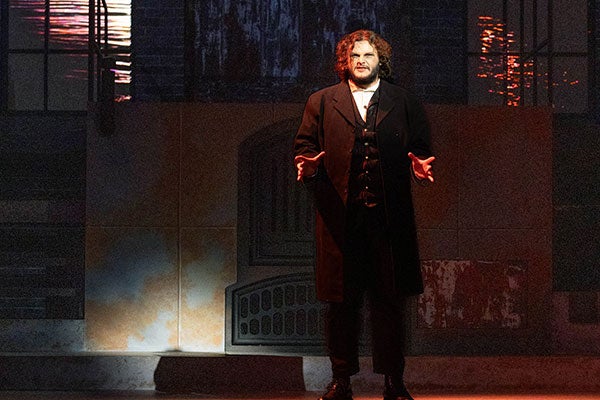 A bearded man wearing a black coat, vest and pants with a white shirt stands facing an audience with his hands and arms opened wide amidst a backdrop of two large windows next to a city street.