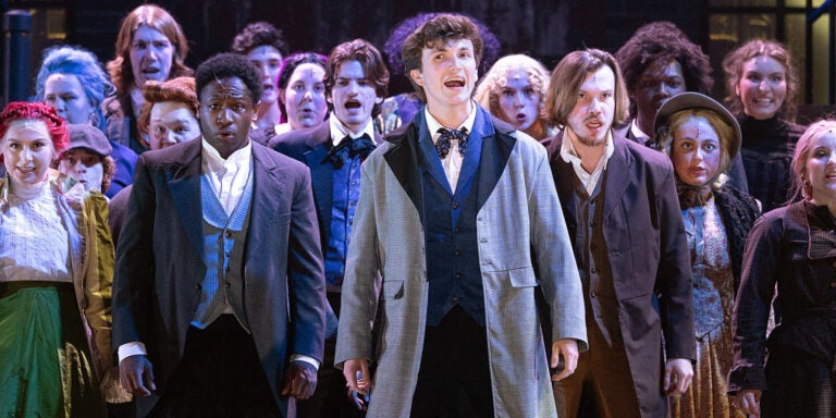 A group of people dressed in clothing that replicates what would have been worn in nineteenth century London stand together on a dark stage during rehearsal for “Sweeney Todd.”