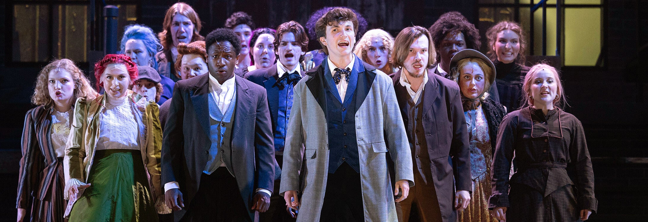 A group of people dressed in clothing that replicates what would have been worn in nineteenth century London stand together on a dark stage during rehearsal for “Sweeney Todd.”