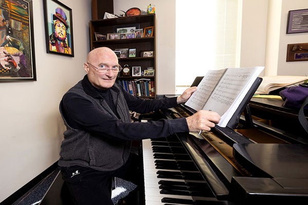 A man sits at a piano in a small office, holding the pages of a musical composition propped on the music stand. Behind him on the wall hang colorful portraits, and stands a bookshelf full of picture frames and books.