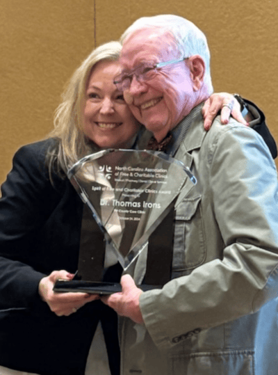 Man in his 70s in olive drab jacket smiling and holding a glass trophy as a middle-aged woman in a black jacket and white blouse puts an arm over his shoulder and squeezes in for a photo taken off to the side.