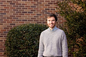 A college-aged man wearing a sweater stands in front of bushes and a brick building.
