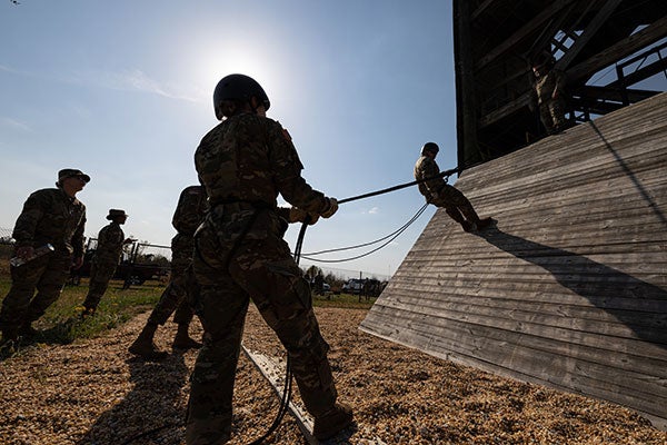 Military cadets cast a shadow while training with ropes and scaling a wooden ramp near the base of a rappelling tower. 