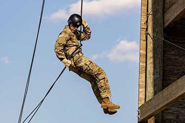 A cadet wearing camouflage and a black helmet lowers himself using ropes off a wooden tower.