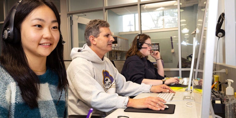 Three people sit in front of computers, two wearing headsets. The closest woman is smiling and writing on a yellow pad of paper.