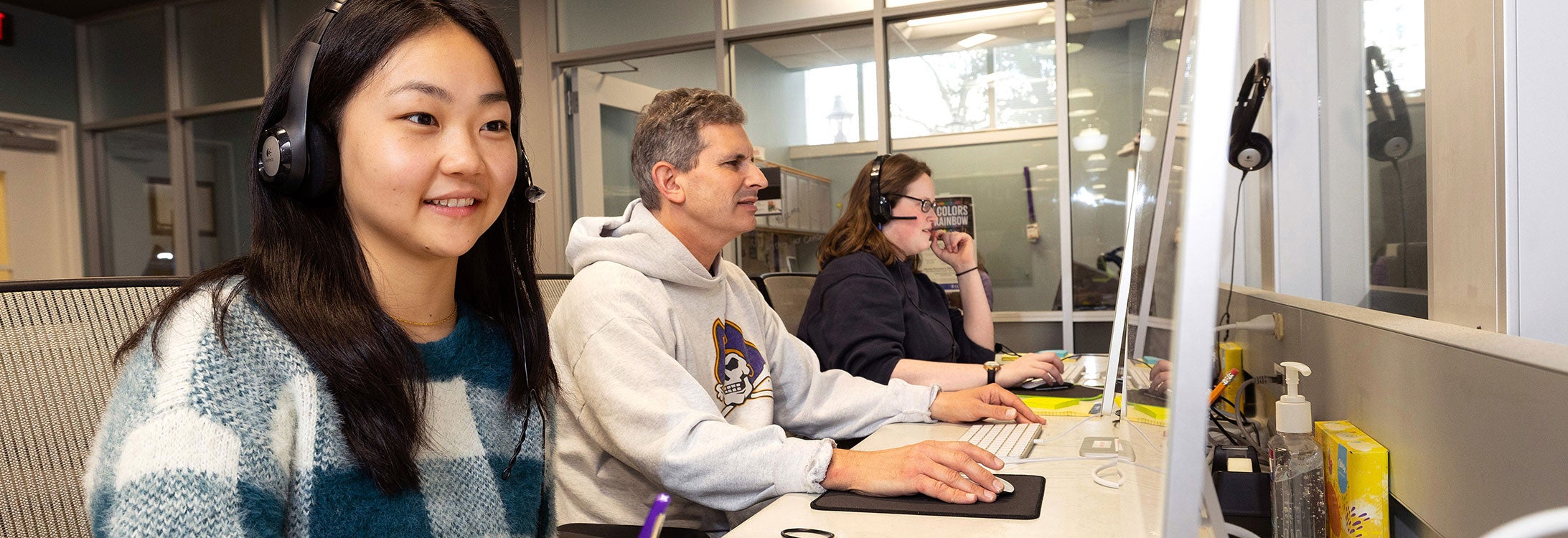 Three people sit in front of computers, two wearing headsets. The closest woman is smiling and writing on a yellow pad of paper.