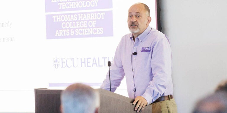 A man in a purple and white shirt stands at a podium and speaks to people who are sitting in front of him.