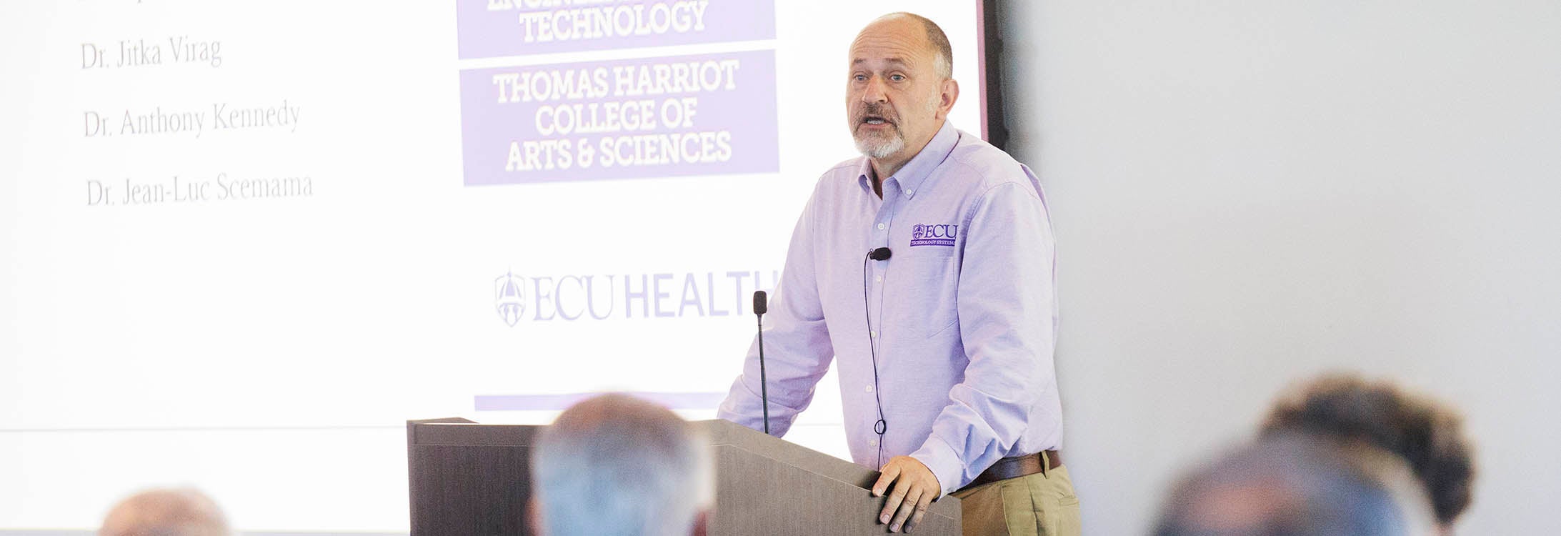 A man in a purple and white shirt stands at a podium and speaks to people who are sitting in front of him.