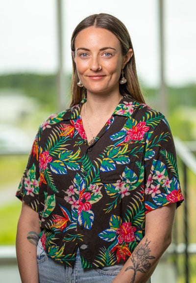 A smiling woman in a black shirt with flowery patterns stands in front of a tall window.