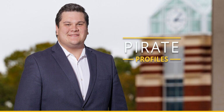 Cole Miller, a male with dark hair and dark eyes smiles at the camera while wearing a white button up shirt and blueish black suit jacket while standing in front of ECU’s Joyner clocktower.