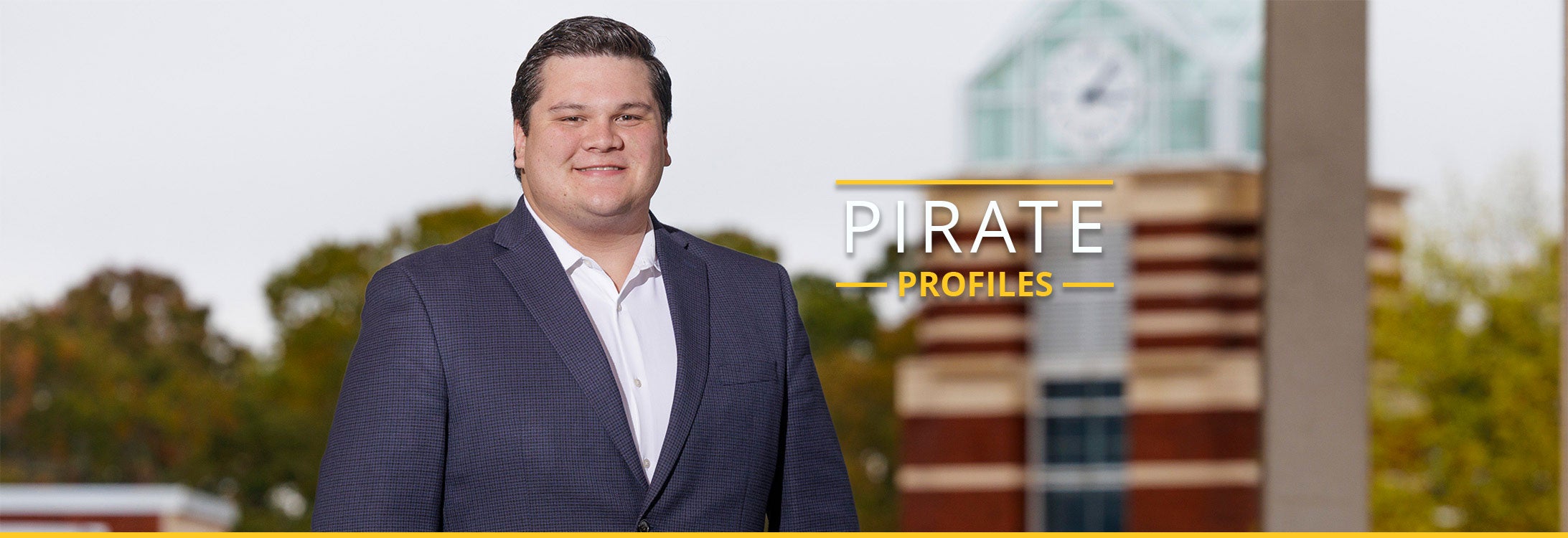 Cole Miller, a male with dark hair and dark eyes smiles at the camera while wearing a white button up shirt and blueish black suit jacket while standing in front of ECU’s Joyner clocktower.