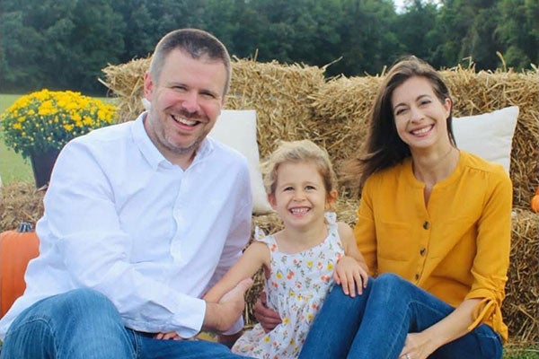 A family consisting of a man with short, blondish hair, a woman with long, dark hair and a young girl with blonde, curly hair smile and pose for a family photo in an open field during autumn.