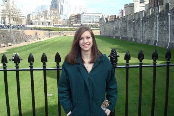 A young, brunette Laurel in a green pea coat smiling and standing in front of a large, black iron gate.