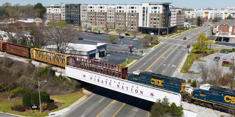 An aerial photo looking over Greenville’s cityscape with a train crossing the Pirate Nation overpass rails in the foreground and housing and businesses in the background.