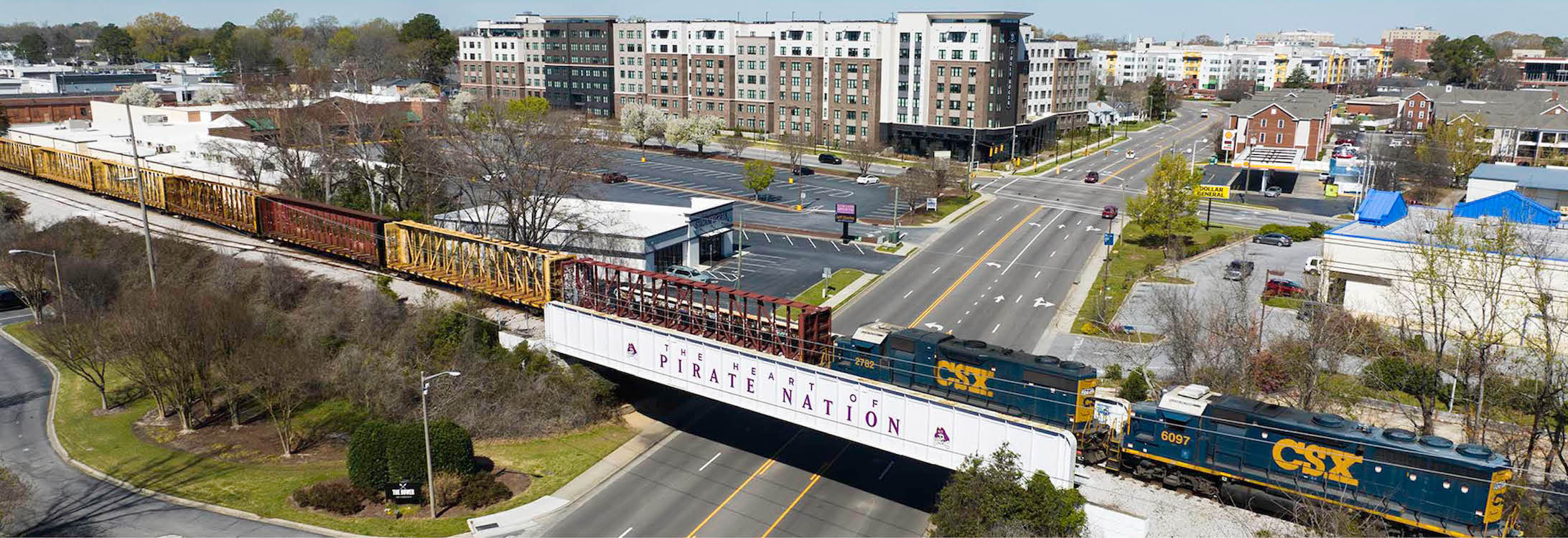 An aerial photo looking over Greenville’s cityscape with a train crossing the Pirate Nation overpass rails in the foreground and housing and businesses in the background.