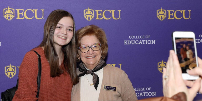 A young woman in a brown sweater smiles as she stands next to an older woman with a black scarf and brown sweater to get their picture made against a purple background. At right, a person holds a cell phone up to take the picture.