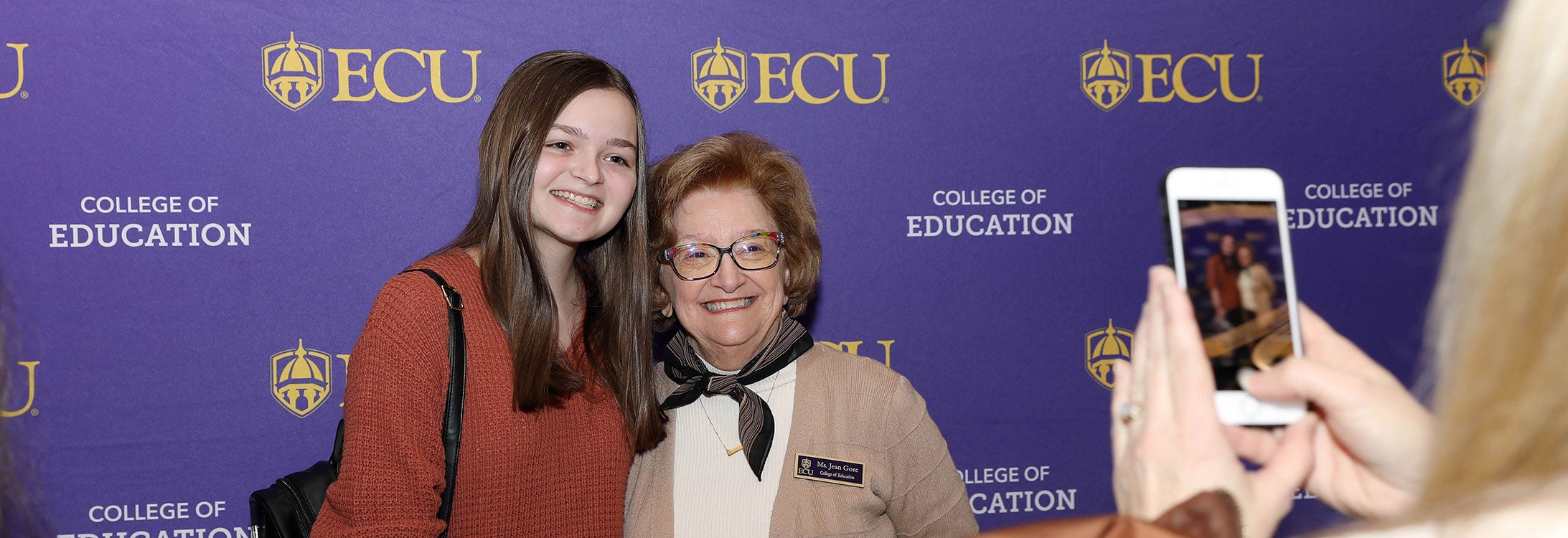 A young woman in a brown sweater smiles as she stands next to an older woman with a black scarf and brown sweater to get their picture made against a purple background. At right, a person holds a cell phone up to take the picture.