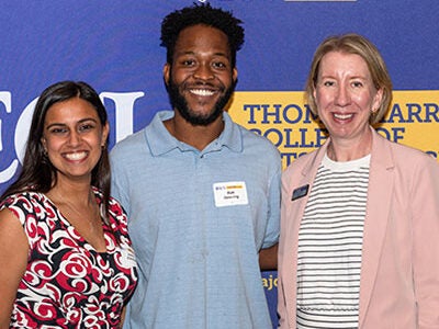 Joanna Kirkendoll and Matt Downing stand to the left of Dean Allison Danell. All three people are smiling and standing in front of a purple, gold, and white photo backdrop with the Harriot College logo.(Photos by Rob Taylor Photography & Design)