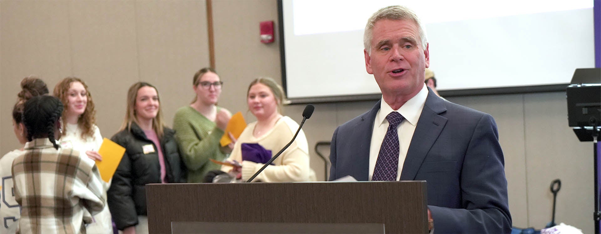 A man wearing a suit and tie stands at a podium and speaks while six young students stand in the background.