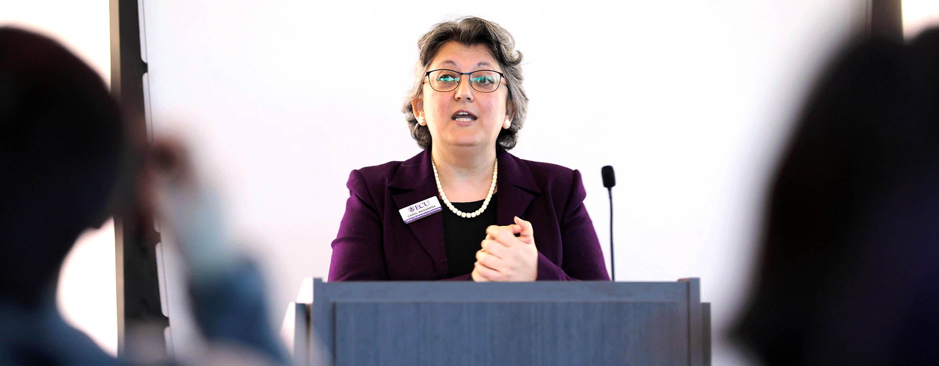 A woman in a purple jacket clasps her hands as she stands at a podium and speaks.