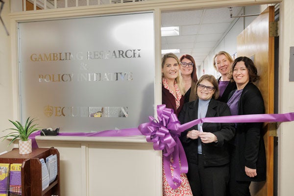 A woman with glasses holds a pair of scissors and is about to cut a ribbon. She is joined by a woman in a red dress to her left, a woman with glasses over her right shoulder, a blonde woman in a purple shirt over her left shoulder and a dark-haired woman to her left.