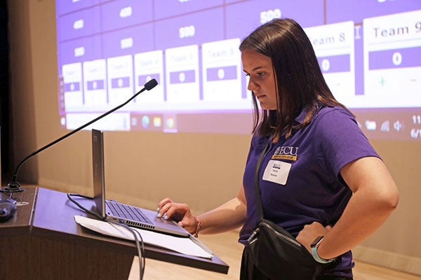 A college age woman wearing a purple East Carolina University polo shirt stands at a podium in a large conference room. 