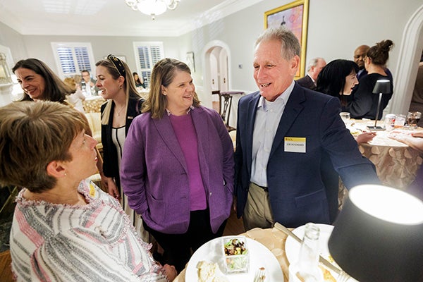 A woman at center in a purple jacket smiles as she listens to a man in a blue sport coat talk while others stand and talk in the background.