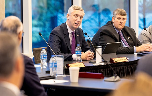 ): A man in a suit with a purple tie sits at a table and speaks surrounded by other people sitting at tables.