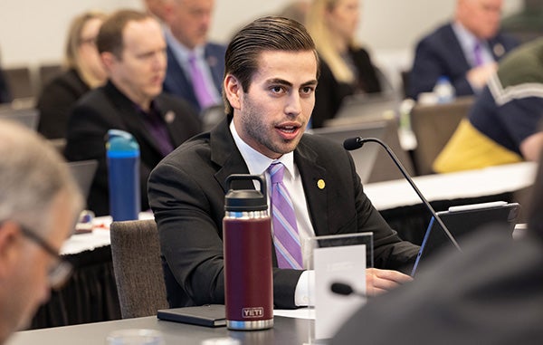 A young man in a suit sits and a table and speaks surrounded by other people sitting at tables.