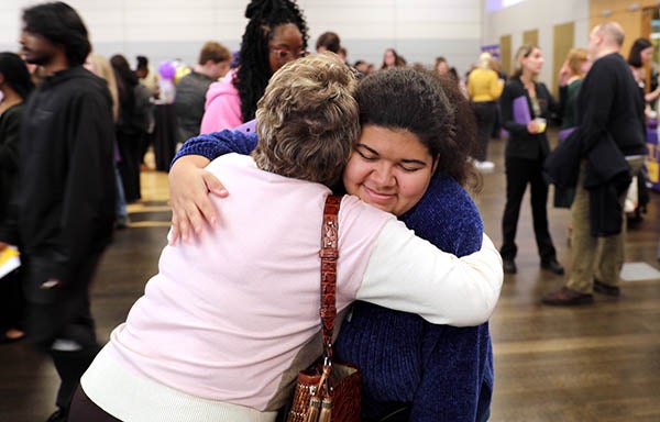 A young woman wearing a blue sweater hugs an older woman wearing a white shirt and pink sweater.