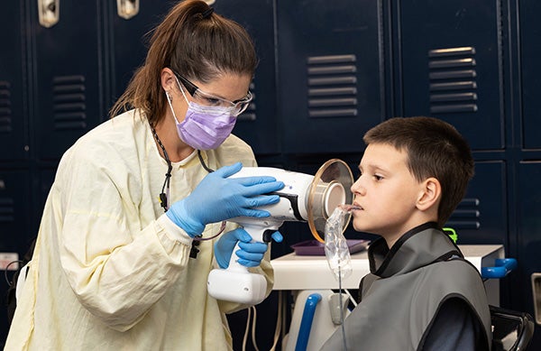 A young boy wearing a protective smock sits straight in a folding chair while a hygienist holds up x mobile x-ray machine to the right side of his face.