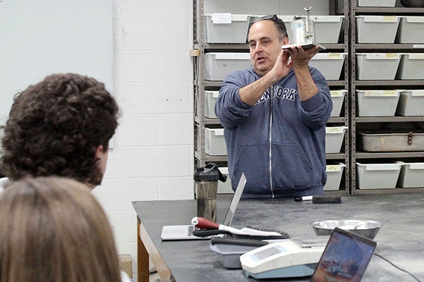 A man in a gray sweatshirt holds up a cylinder to show students sitting at a lab table.