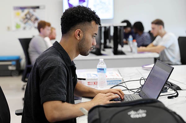 ): A college student in a black shirt types on a laptop computer while sitting at a long desk. Other students in the background type on computers as well.