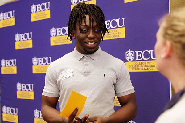 : A young man wearing a gray polo shirt smiles as he speaks to a woman standing at right.