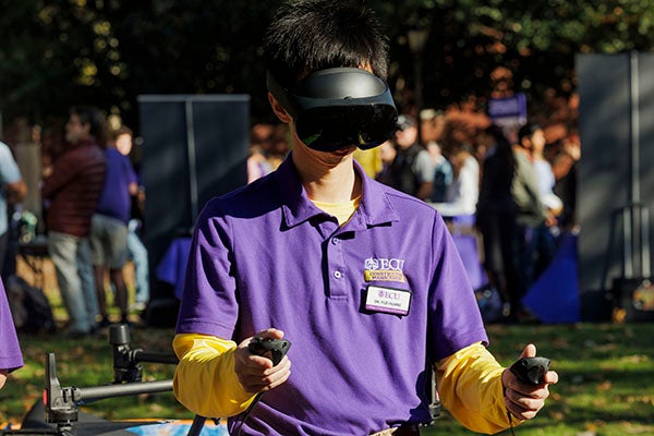 A man in a purple shirt wears a virtual reality headset and holds a controller in each hand.