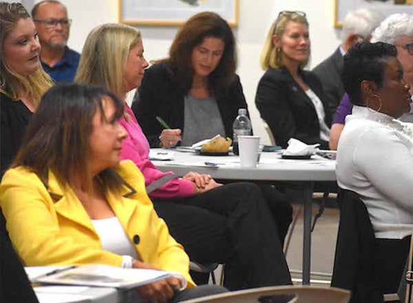 Several women sit around round tables in business casual while listening to a speaker. 