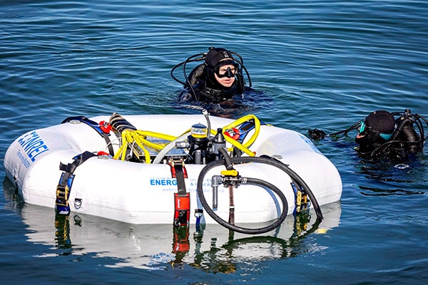 Two divers in scuba gear swim next to an inflatable raft containing equipment to generate electricity from wave energy.