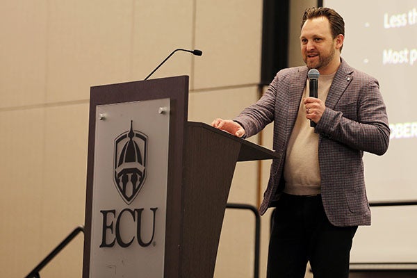 ): A man in a brown jacket holds a microphone as he stands next to a podium and speaks.