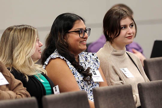 Three young women, one at left in a black sweater, one in the middle with a flowered shirt and one at right in a brown sweater, sit in chairs and listen to a presentation. The woman in the middle is smiling.