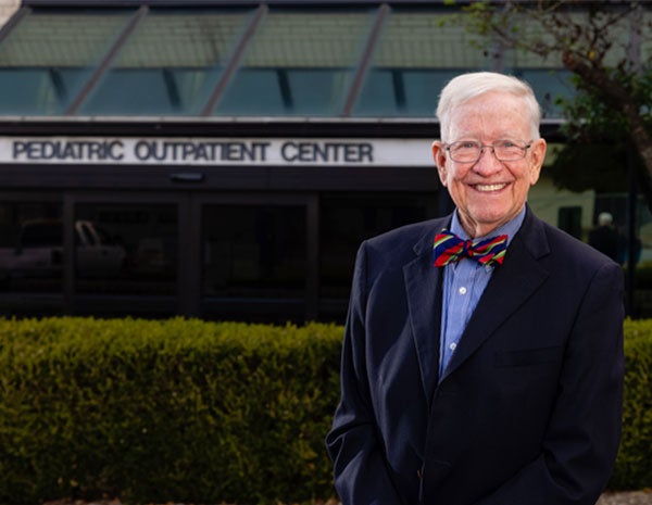 Man in his 70s in a colorful bowtie and navy blue jacket stands smiling outside a Pediatric Outpatient Center.