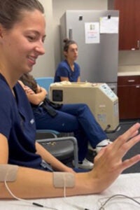 A woman with dark hair wearing scrubs is seated with wires attached to her right arm.