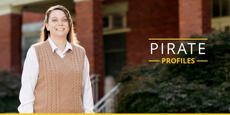 A woman in a white shirt and brown sweater stands in front of a building and bushes.