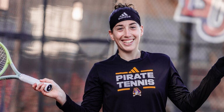 A smiling young woman in a black Pirate Tennis long sleeve T-shirt holds a tennis racket in her right hand.