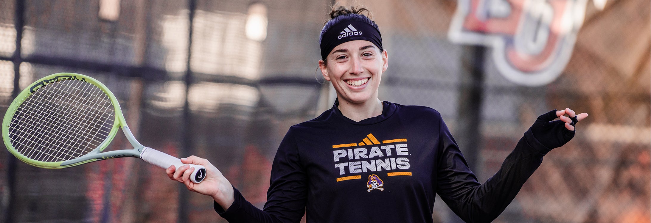 A smiling young woman in a black Pirate Tennis long sleeve T-shirt holds a tennis racket in her right hand.