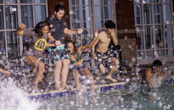 A young woman holds hands with a man as they begin to jump into a swimming pool. To their right, another young woman and man jump into the pool. 